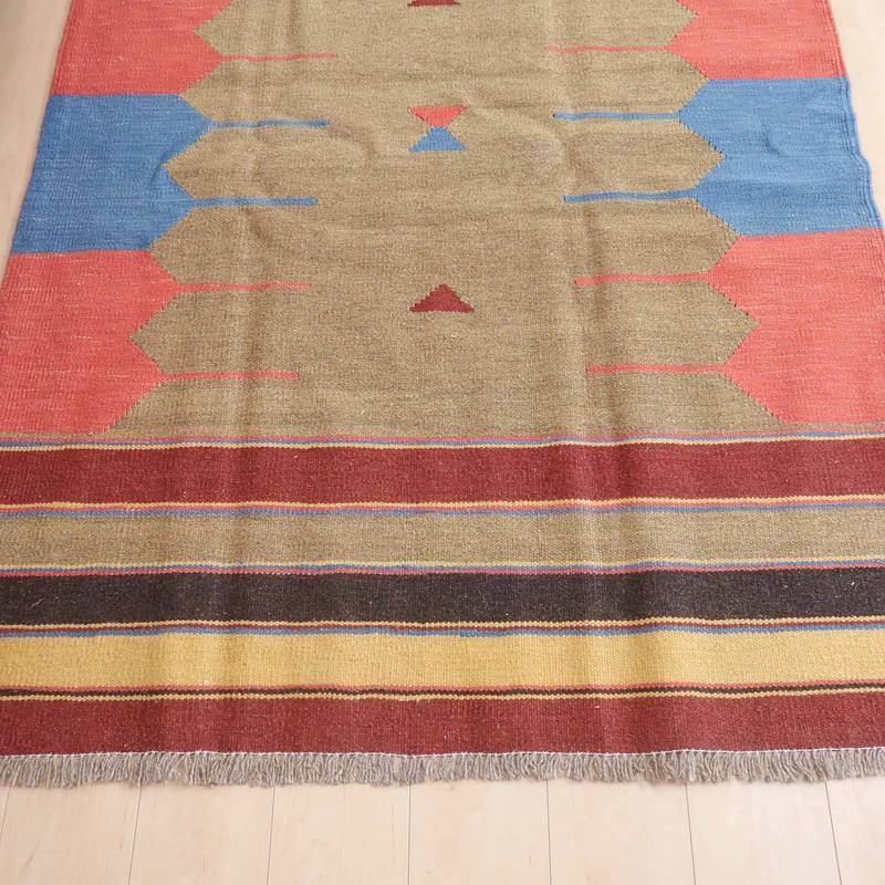 ベージュを基調に赤と青の幾何学模様が織り込まれた手織りのギャッベラグが、ペルシャ絨毯専門店の通販で床に広がっている。