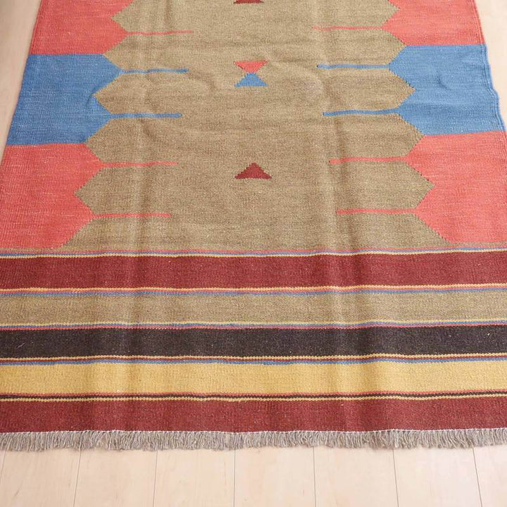 ベージュを基調に赤と青の幾何学模様が織り込まれた手織りのギャッベラグが、ペルシャ絨毯専門店の通販で床に広がっている。