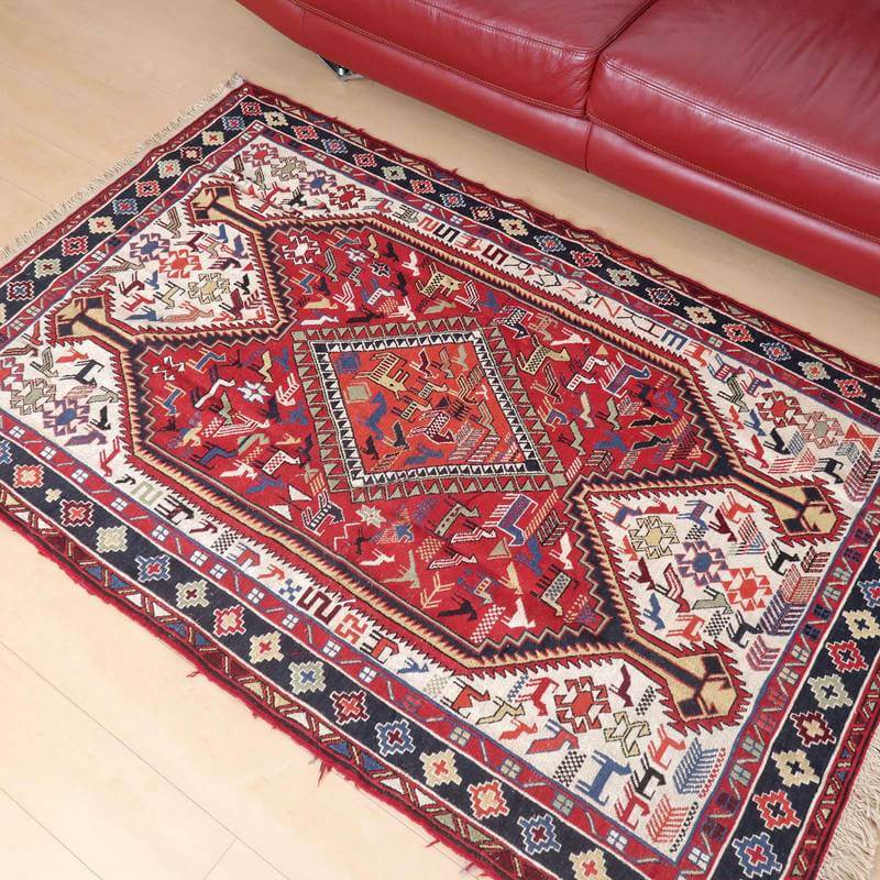 赤いソファの前に敷かれた、鮮やかな色彩と複雑な模様が美しい手織りのペルシャ絨毯ギャッベラグが床に映えている。