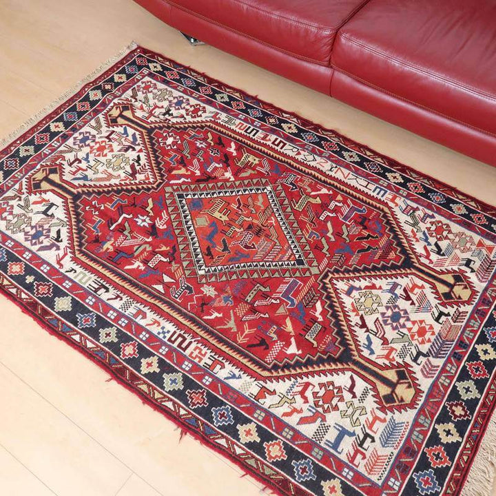赤いソファの前に敷かれた、鮮やかな色彩と複雑な模様が美しい手織りのペルシャ絨毯ギャッベラグが床に映えている。