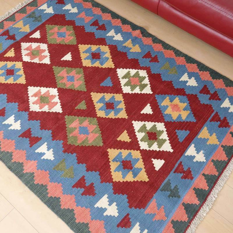 カラフルな幾何学模様が特徴の手織りギャッベラグが、赤いソファのそばに敷かれている様子が見える。