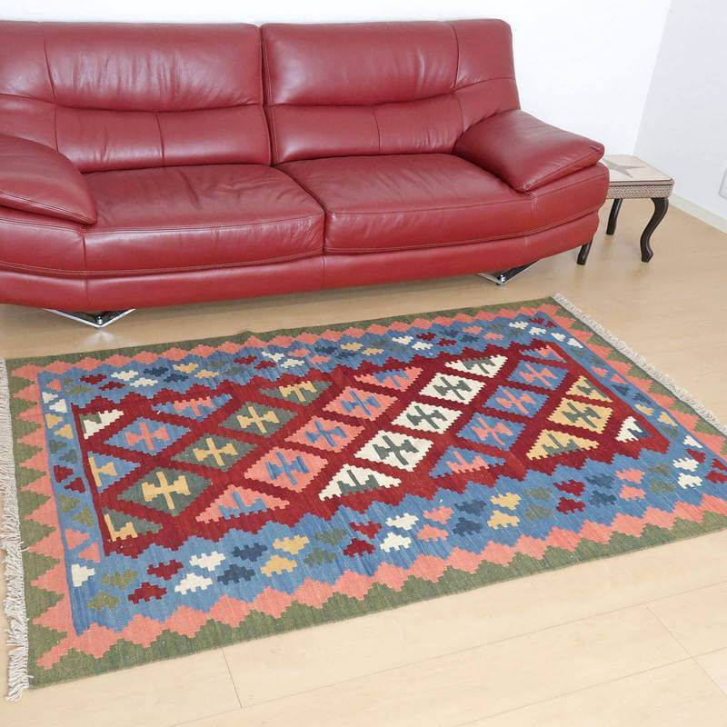 赤いソファの前に敷かれたカラフルな模様のギャッベラグが、ペルシャ絨毯専門店の手織りウール製品として通販で見える。