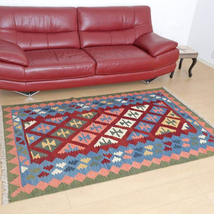 赤いソファの前に敷かれたカラフルな模様のギャッベラグが、ペルシャ絨毯専門店の手織りウール製品として通販で見える。
