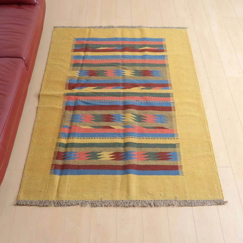 キリム・シラーズ/カシュガイ 189×128cm(KL-406)は、黄色を基調にカラフルなストライプ模様が施された手織りのウールラグがリビングの床に敷かれている。
