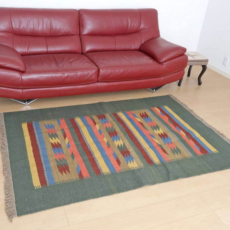 赤いソファの前に、カラフルな模様が特徴の手織りギャッベラグが敷かれており、ウール素材の温かみがリビングに広がっている。