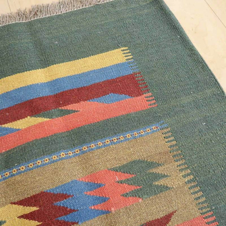 緑を基調に赤と青の幾何学模様が鮮やかに手織りされたギャッベのラグが床に敷かれ、ペルシャ絨毯の伝統を感じさせます。