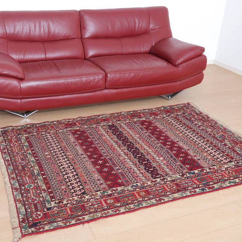 赤いレザーソファの前に、伝統的なペルシャ絨毯の模様が美しい手織りのウール製ギャッベラグが敷かれている。
