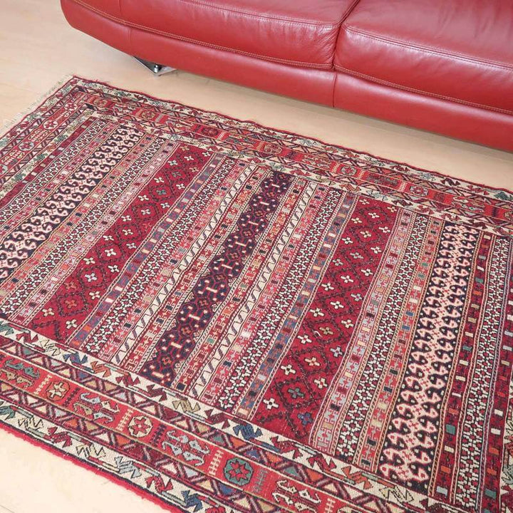 手織りのギャッベラグがリビングの赤いソファ前に敷かれ、ペルシャ絨毯の伝統的な模様とウールの質感が床に映えている。