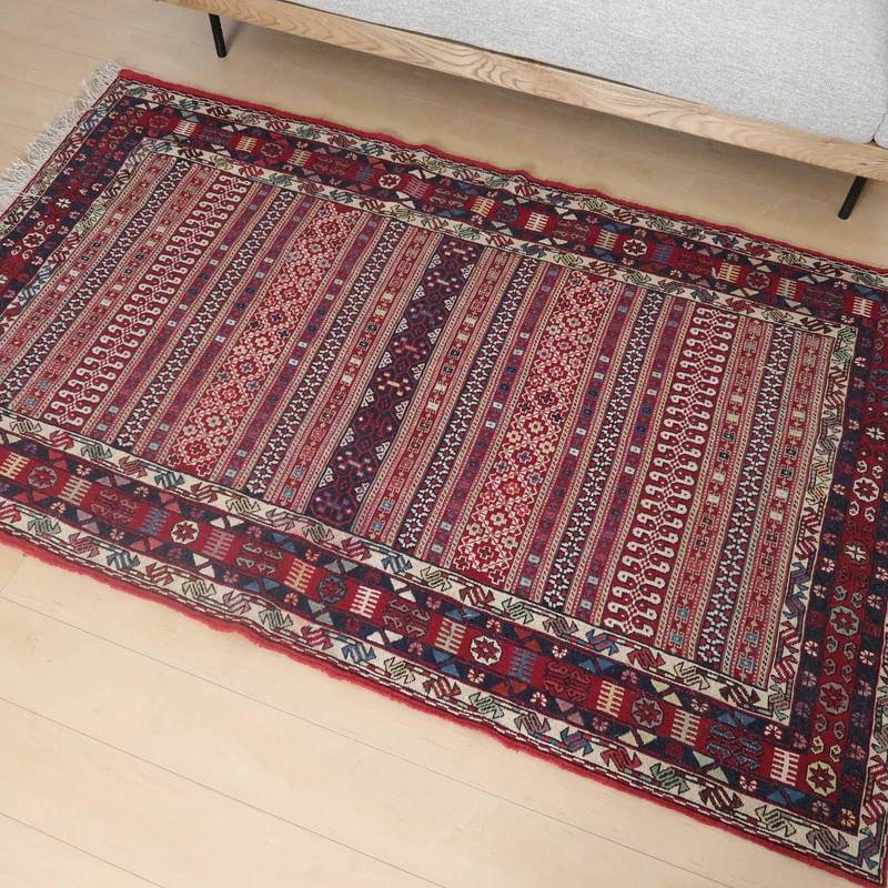 赤や青の色合いが美しい手織りのギャッベラグが、ウール素材で丁寧に仕上げられ、木製家具が置かれたリビングの床に映えています。