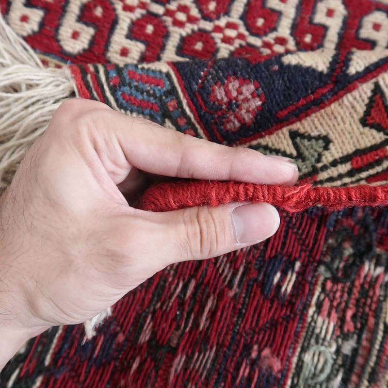 手織りのウールで丁寧に作られたペルシャ絨毯の縁を持ち上げ、厚みと織りの密度を確認する様子が見える。