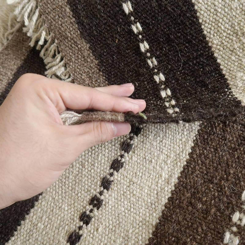 茶色とベージュのストライプが並ぶ手織りのギャッベラグが、ウールの質感と織りの密度を指でめくっている様子が見える。