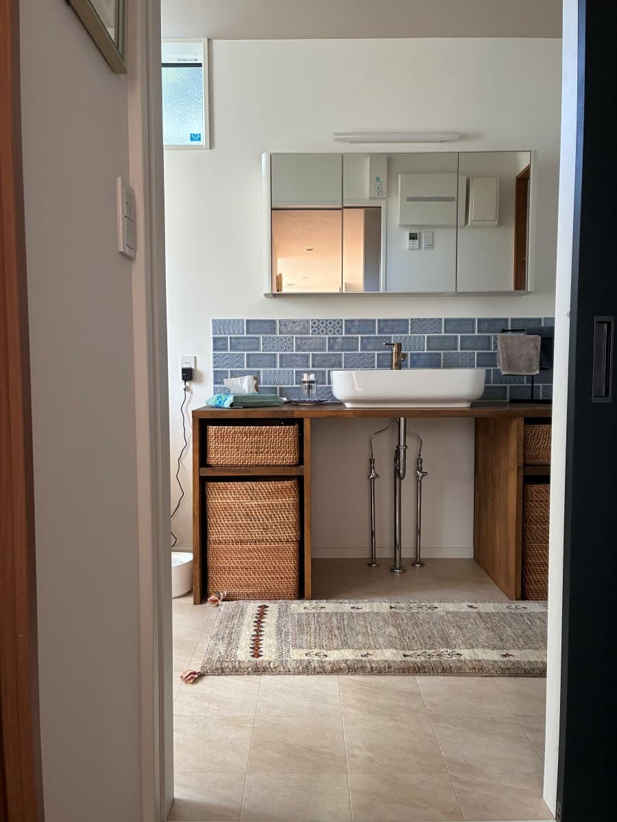 洗面台横の通路に、ラタン収納と木のカウンターに馴染む細長いベージュのギャッベをコーディネート