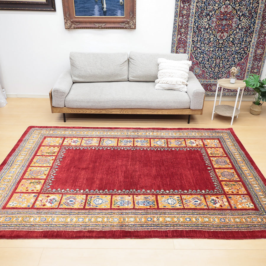 赤を基調とした伝統的な模様の敷物が床に敷かれ、後ろにグレーのソファとクッションが置かれている部屋。