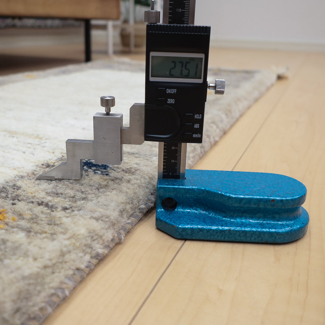 カーペットの厚さをデジタルノギスで測定している様子、床は木製で背景に家具がある。