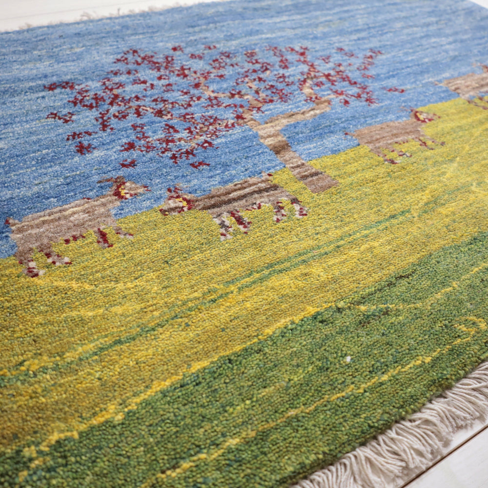 青空と草原を背景に、動物と木が丁寧に手織りされたウール製のペルシャ絨毯ギャッベが床に広がっている。