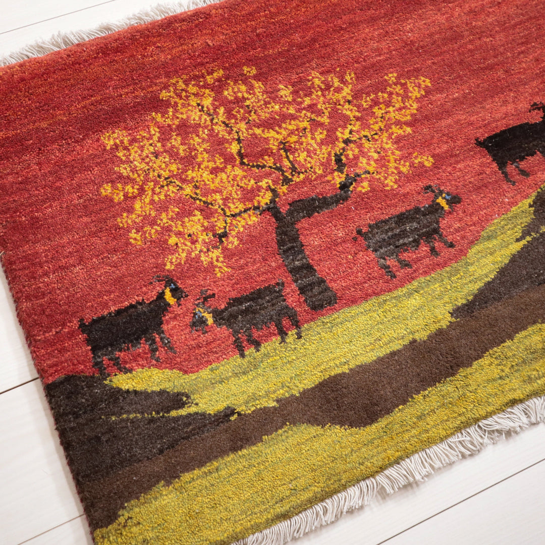 燃えるような赤い背景に黒いヤギと黄色の木が描かれたウール製の手織りギャッベラグが床に広がり、その織り目が際立っている。