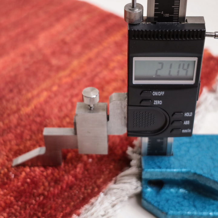 赤い手織りウールのギャッベラグが計測器で厚みを測定され、織りの緻密さが際立っている。