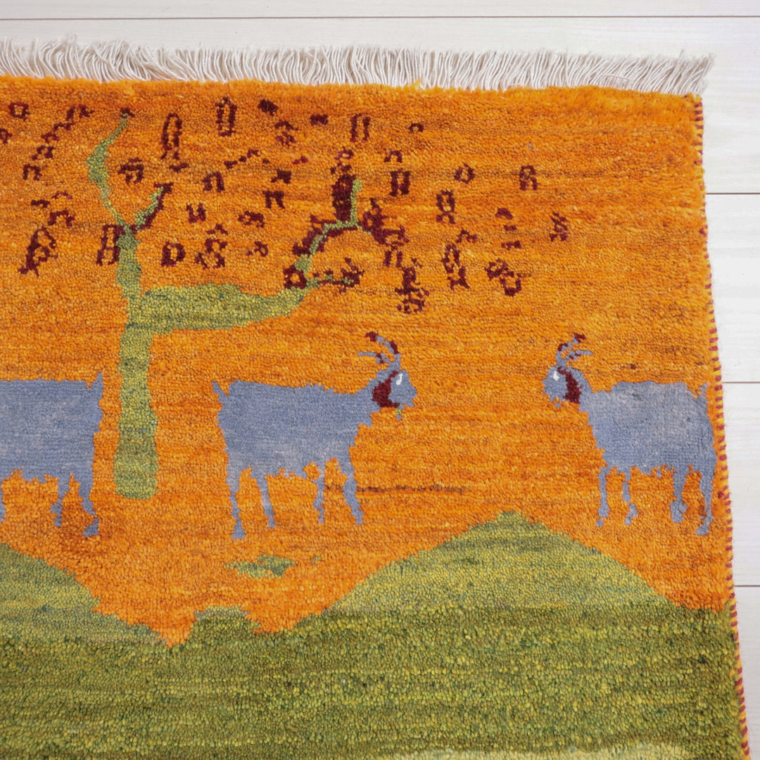 オレンジと緑の鮮やかな色合いで手織りされたギャッベラグには、ペルシャ絨毯専門店で通販可能な山羊と木のデザインが生き生きと描かれている。