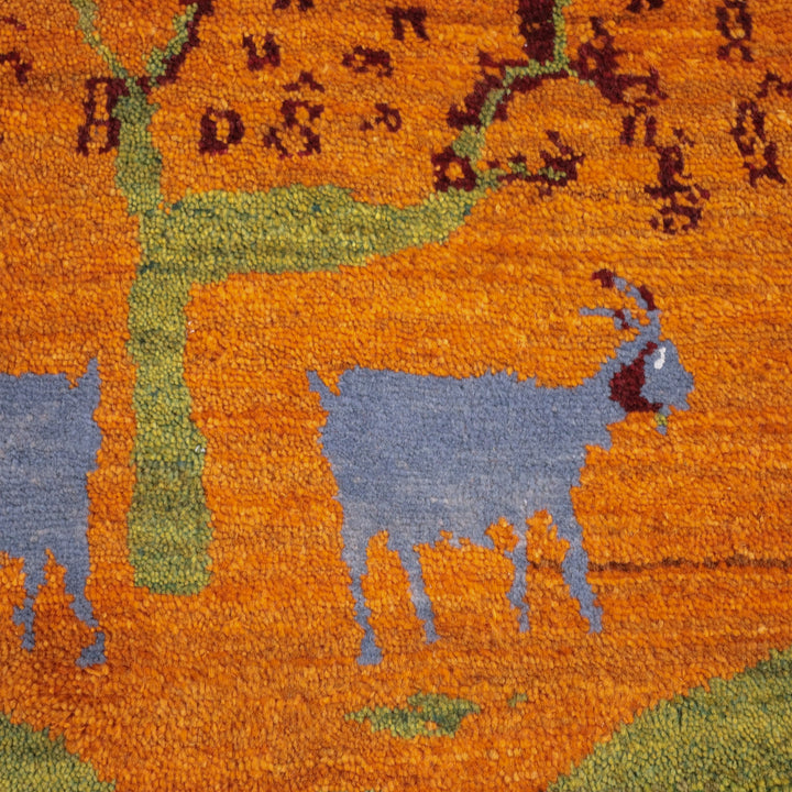 オレンジとブルーの羊が描かれた手織りのギャッベラグが、豊かな自然を背景にしたペルシャ絨毯専門店の通販アイテムとして床に広がっている。
