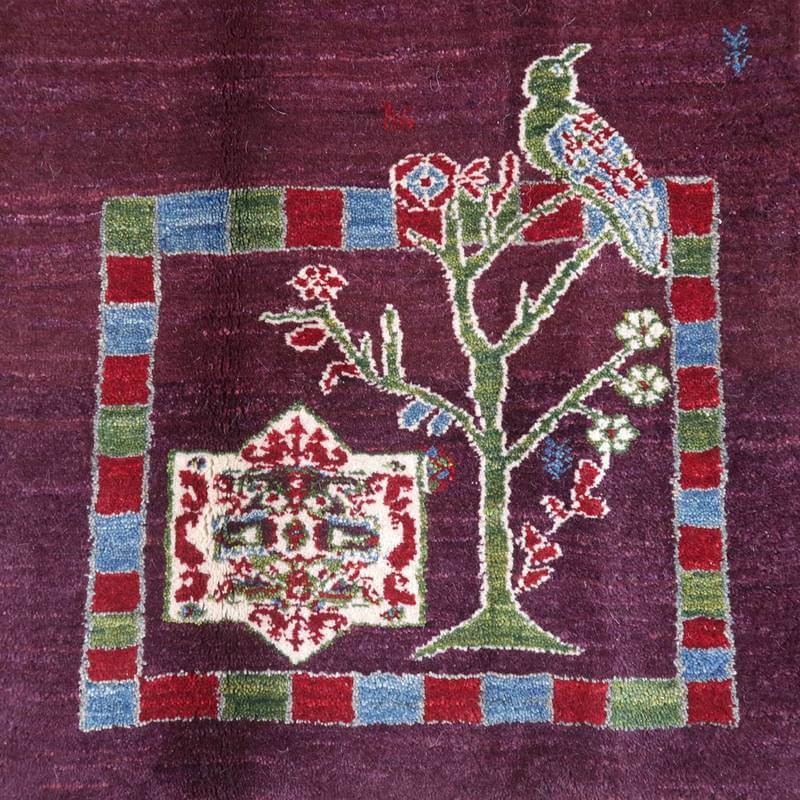 紫の背景に色鮮やかな鳥と木のモチーフが丁寧に手織りされたギャッベのラグが敷かれている。