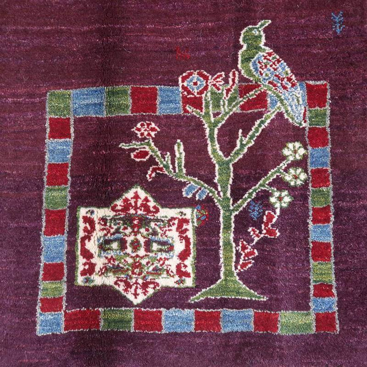 紫の背景に色鮮やかな鳥と木のモチーフが丁寧に手織りされたギャッベのラグが敷かれている。