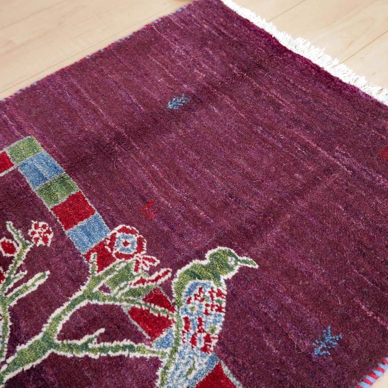 紫色の濃厚な手織りウールのギャッベラグに、ペルシャ絨毯の専門店ならではの鳥と花の模様が丁寧に織り込まれている様子が見える。