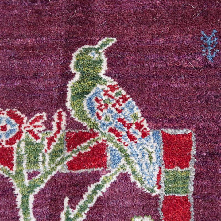 緻密に手織りされたペルシャ絨毯の一部で、鮮やかな鳥のデザインが深い赤の背景に映えるウール製ギャッベラグが見える。