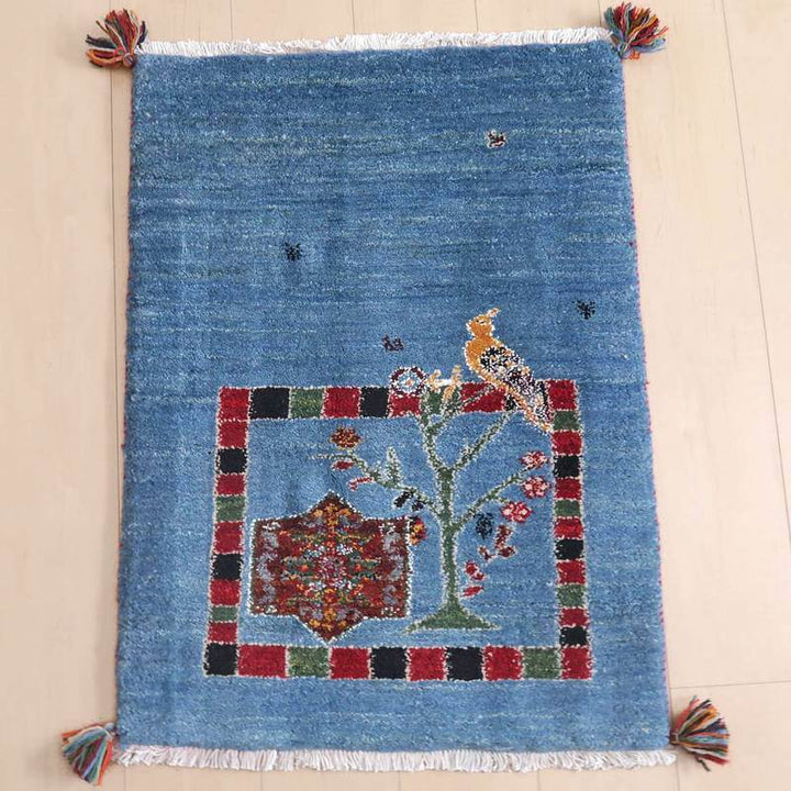 ギャッベ／ルリバフ 81×61cm（HSS-73157）は、青い背景に鳥と花のモチーフが鮮やかに描かれた手織りのウール製ギャッベラグが床に敷かれている様子が見える。