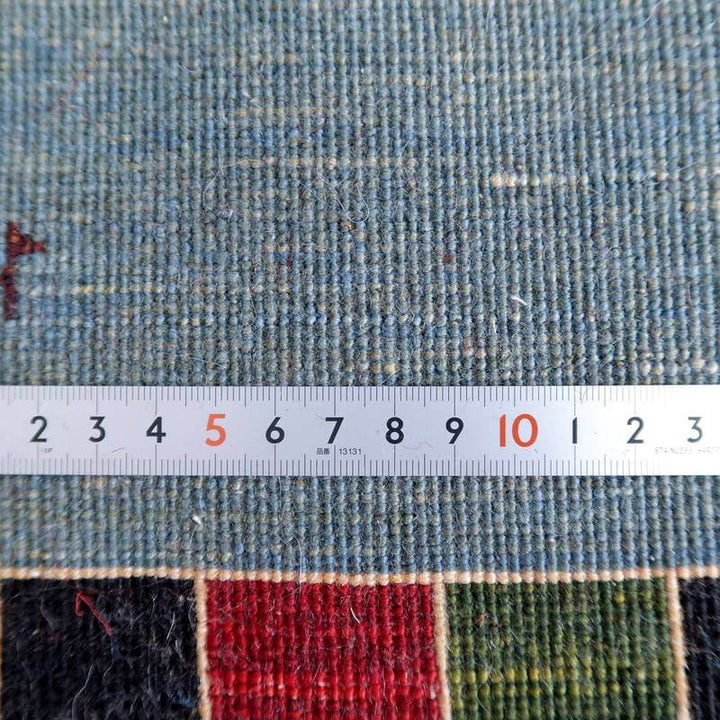 青い手織りのウール製ギャッベラグの織り目が精巧に見え、定規でその密度が測定されている様子が映し出されています。