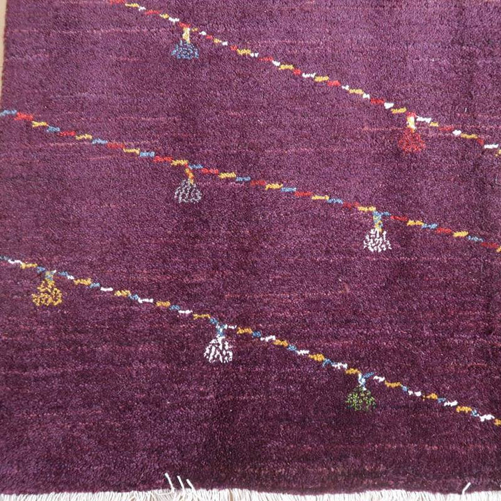 深みのある紫色のウール製ギャッベラグが、丁寧に手織りされたペルシャ絨毯のデザインを背景に、色とりどりの模様が浮かび上がっている。