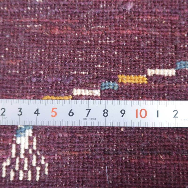 紫色のウール素材が丁寧に手織りされたギャッベラグの繊細な織り目が、定規を用いてその密度が計測されている様子。
