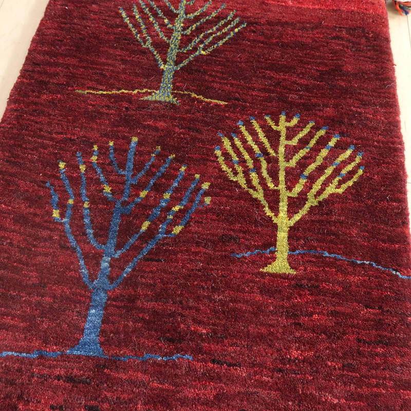 深みのある赤色のウール素材に、手織りで描かれたカラフルな木の模様が印象的なギャッベラグが見える。