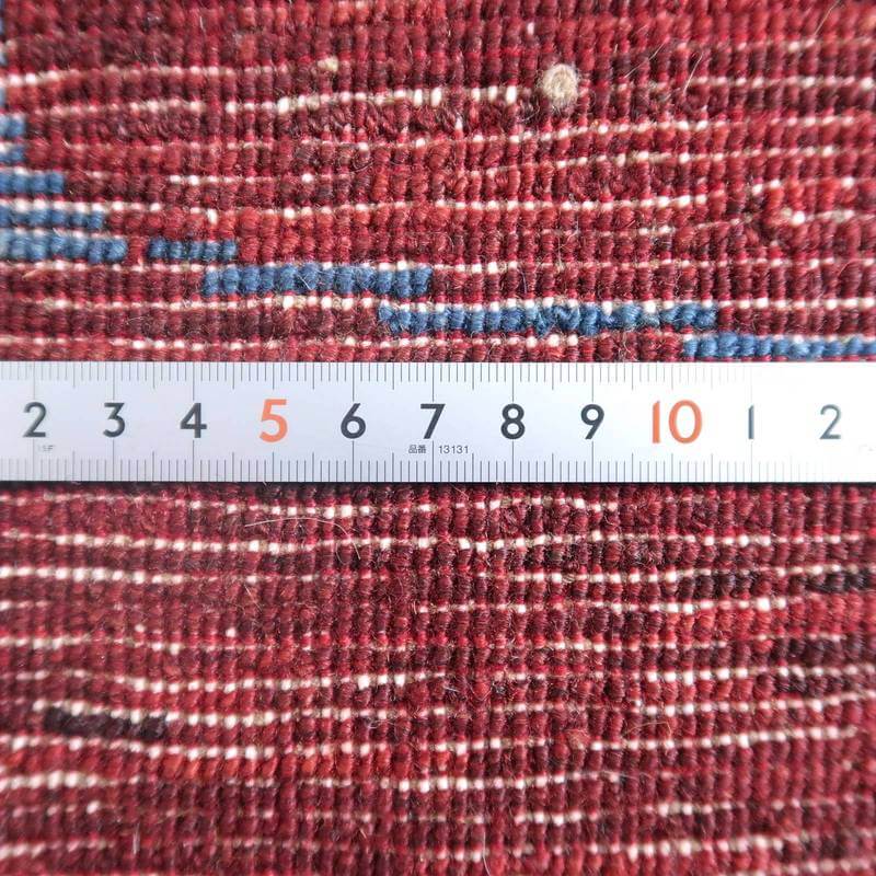 手織りのウール素材が温かみを感じさせるギャッベラグ、ペルシャ絨毯専門店通販で販売中の一枚に、赤い織り目の密度が定規とともに際立っている。
