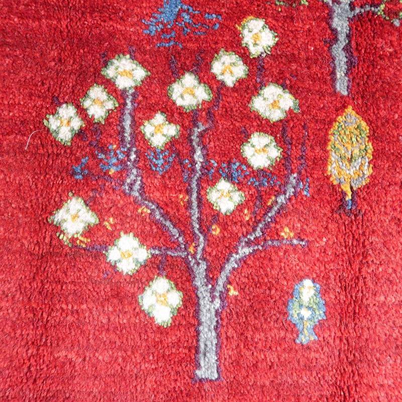 鮮やかな赤色の手織りギャッベラグに、白い花が咲く木が美しく描かれ、ペルシャ絨毯専門店の通販で購入可能な一品です。