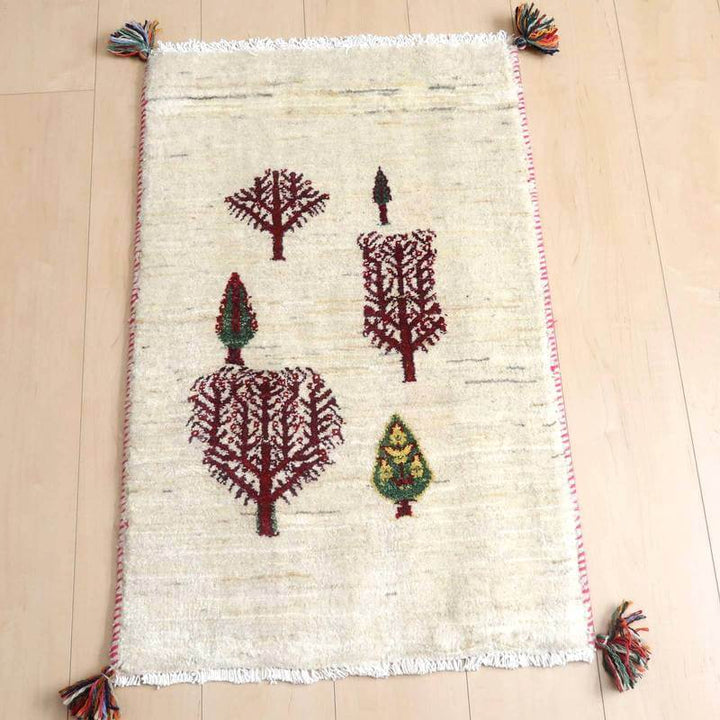 ギャッベ／ルリバフ 85×55cm（HSS-71543）は、白地に赤や緑の木のモチーフが丁寧に手織りされたギャッベのラグが床に敷かれている。