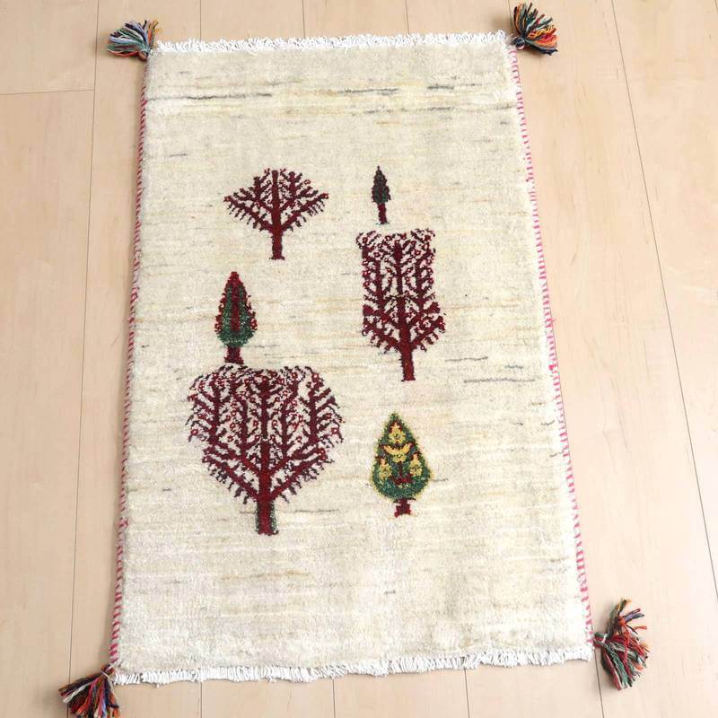 ギャッベ／ルリバフ 85×55cm（HSS-71543）は、白地に赤や緑の木のモチーフが丁寧に手織りされたギャッベのラグが床に敷かれている。