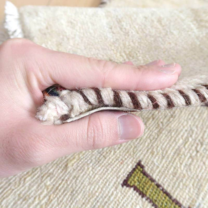 手織りのギャッベラグの縁を指でつまんで厚みを確認している様子が見え、ウールの質感とペルシャ絨毯の魅力が際立っている。