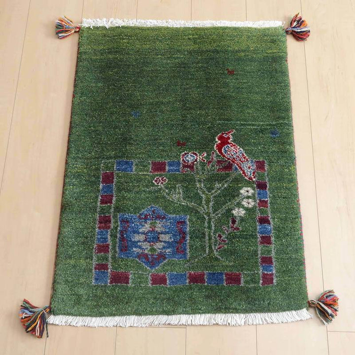 ギャッベ／ルリバフ 78×57cm（HSS-71692）は、緑の背景に鳥や花のデザインが印象的な手織りのウール製ギャッベラグが、明るい木製の床に美しく敷かれている。