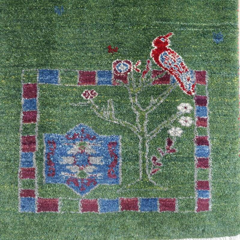 緑の背景に赤い鳥と花の模様が丁寧に織られたこの手織りのギャッベラグは、ウール素材の温かみが感じられるペルシャ絨毯として専門店の通販で見られる。