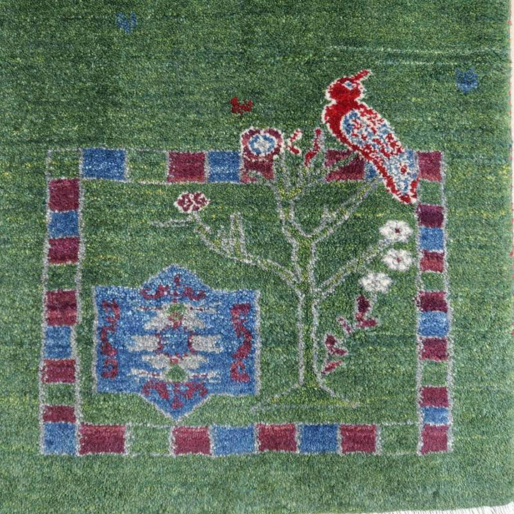 緑の背景に赤い鳥と花の模様が丁寧に織られたこの手織りのギャッベラグは、ウール素材の温かみが感じられるペルシャ絨毯として専門店の通販で見られる。