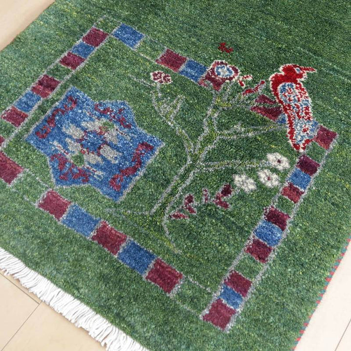 緑豊かな手織りのギャッベラグには、赤い鳥と花の繊細なデザインが施され、ウールの温もりが感じられる。