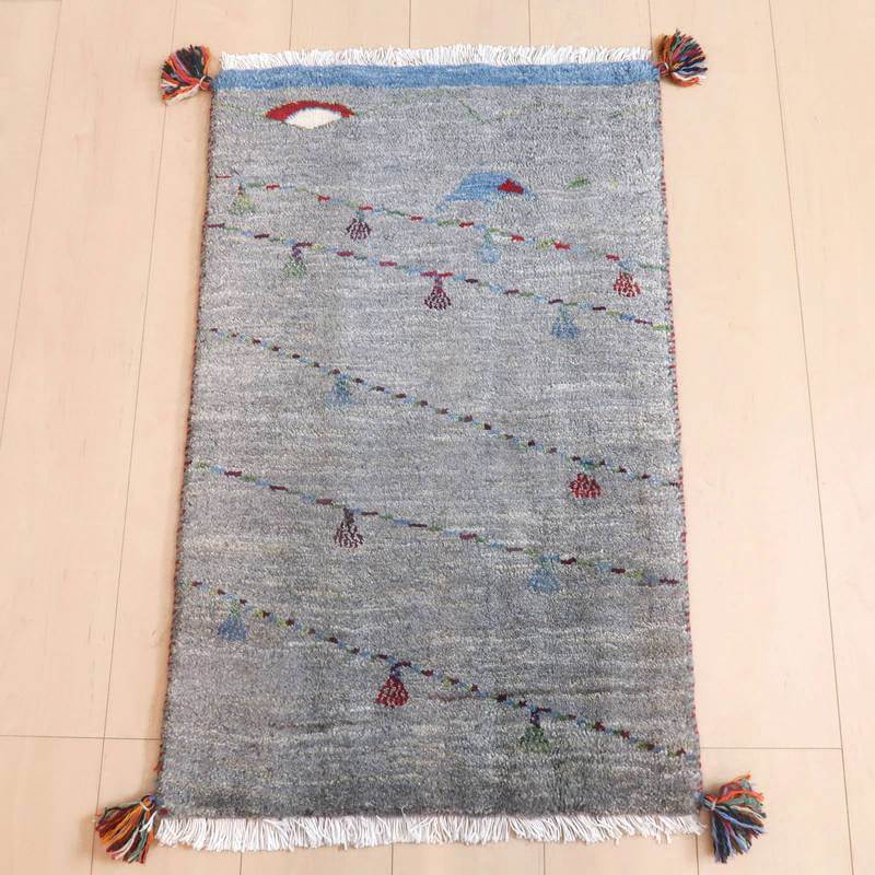 ギャッベ／ルリバフ 86×55cm（HSS-71706）は、手織りのギャッベラグが明るい木目の床に敷かれており、落ち着いたグレートーンにカラフルなモチーフが散りばめられたデザインが映えています。
