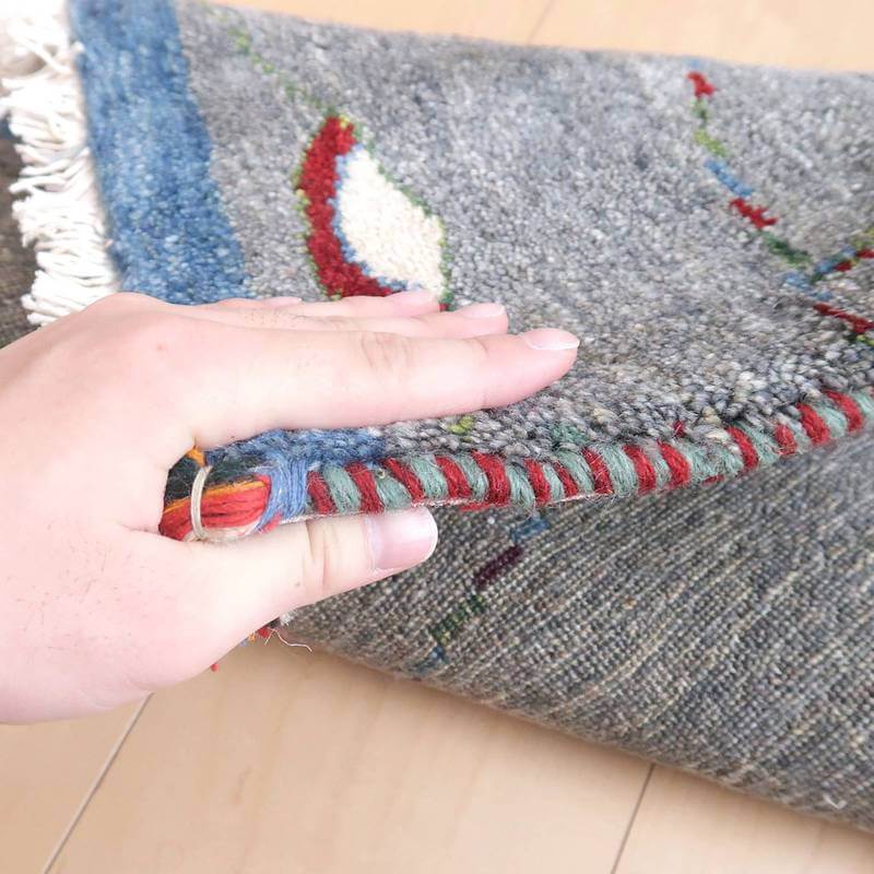 丁寧に手織りされたウールのギャッベラグの厚みが手でめくられ、色鮮やかな縁取りが際立っている。