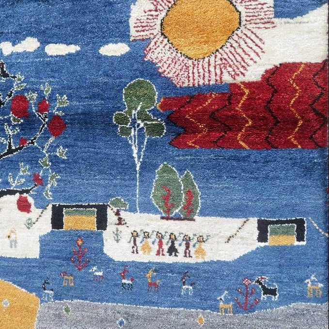 青い背景に赤い実をつけた木や家、太陽が描かれた手織りのウール製ギャッベラグが床に広がり、ペルシャ絨毯専門店の通販で取り扱われています。