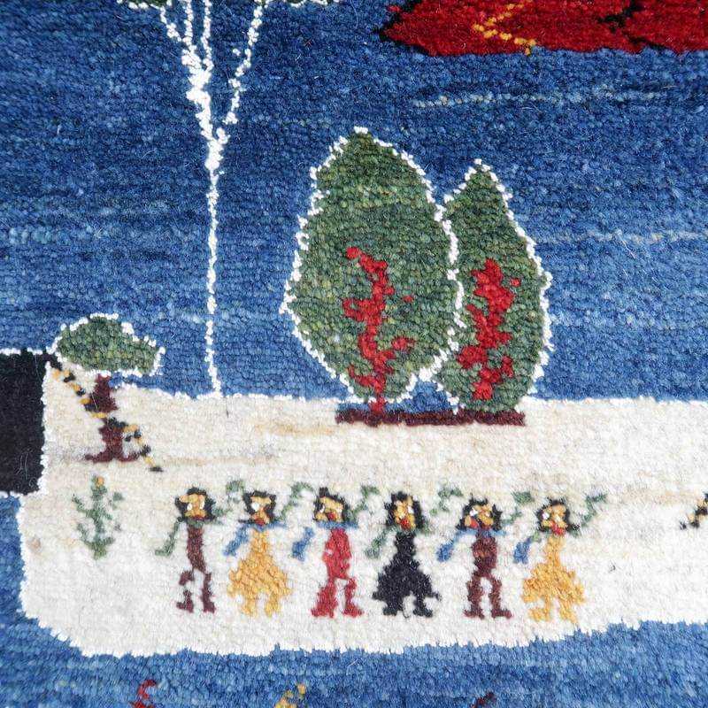 色鮮やかな青と緑の背景に、手織りで表現された人々と木々が描かれたギャッベのラグが見える。