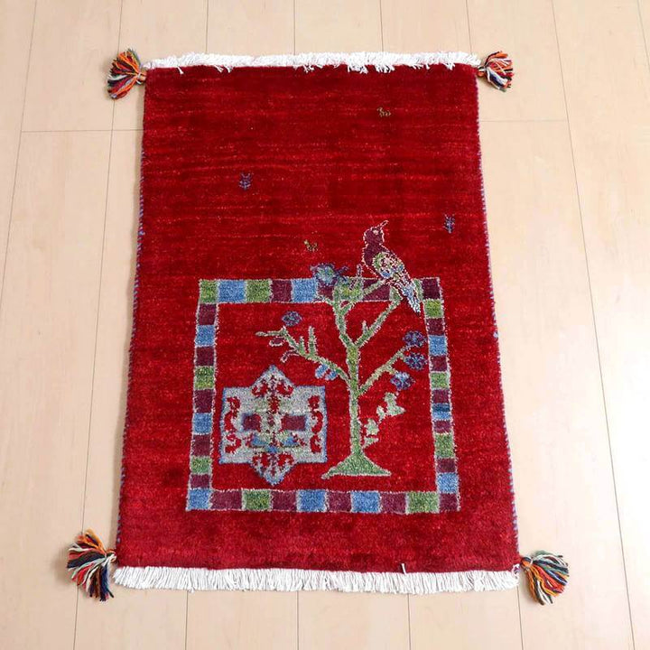 ギャッベ／ルリバフ 81×56cm（HSS-71758）は、赤い背景に色鮮やかな鳥と木のデザインが施された手織りのウール製ギャッベラグが床に敷かれている。