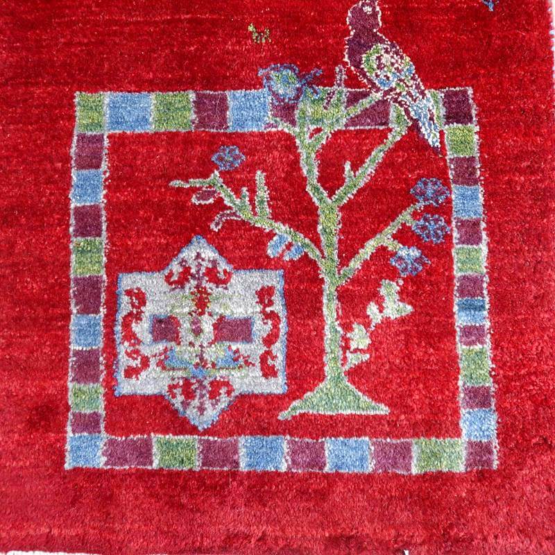 赤い背景に緑と青の鳥や木のモチーフが丁寧に手織りされたウール製ギャッベのラグが見える。