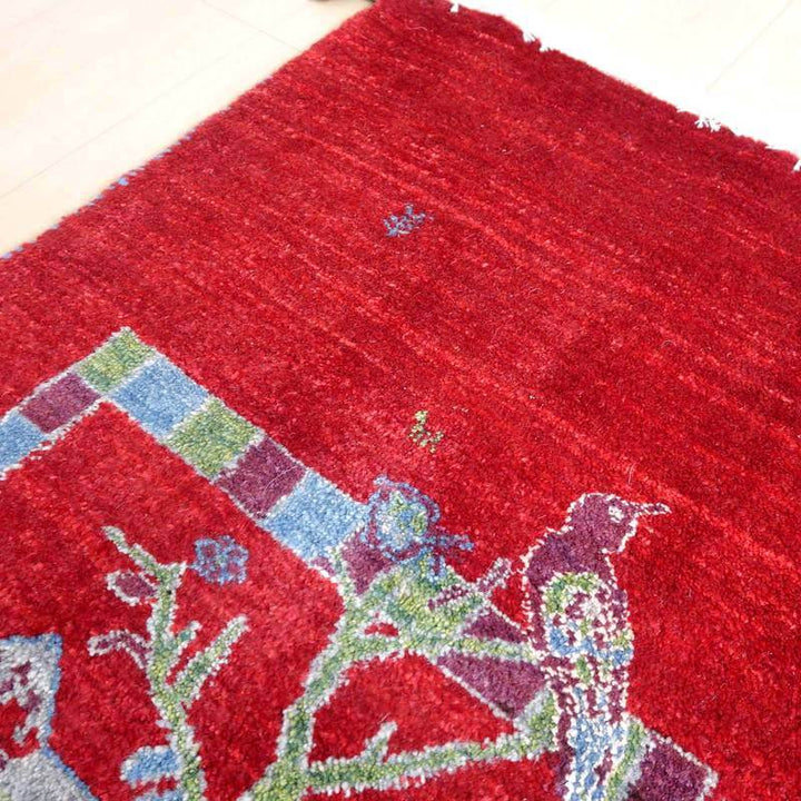 鮮やかな赤い色調のギャッベラグには、鳥と植物の模様が手織りウールで丁寧に表現され、ペルシャ絨毯専門店からの通販で購入可能な一品が床に広がっている。