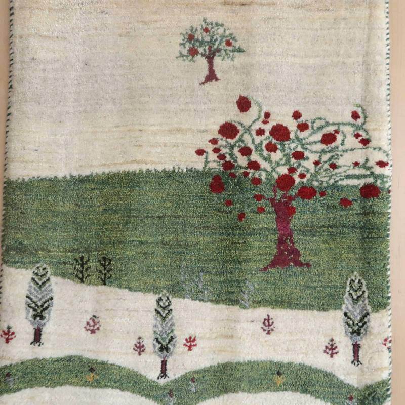 手織りのギャッベラグに描かれたペルシャ絨毯の赤い木々と緑の風景が、ウールの温かみと共に専門店からの通販で手に入る。