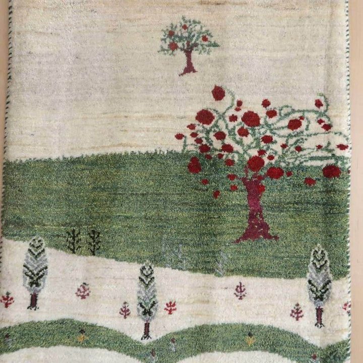 手織りのギャッベラグに描かれたペルシャ絨毯の赤い木々と緑の風景が、ウールの温かみと共に専門店からの通販で手に入る。
